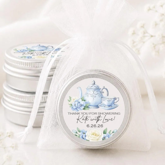 a set of silver tin containers with white labels, each containing a teapot and teacup illustration. The labels are white and have a floral design. The containers are placed on a white surface, and there are some white flowers in the background.
