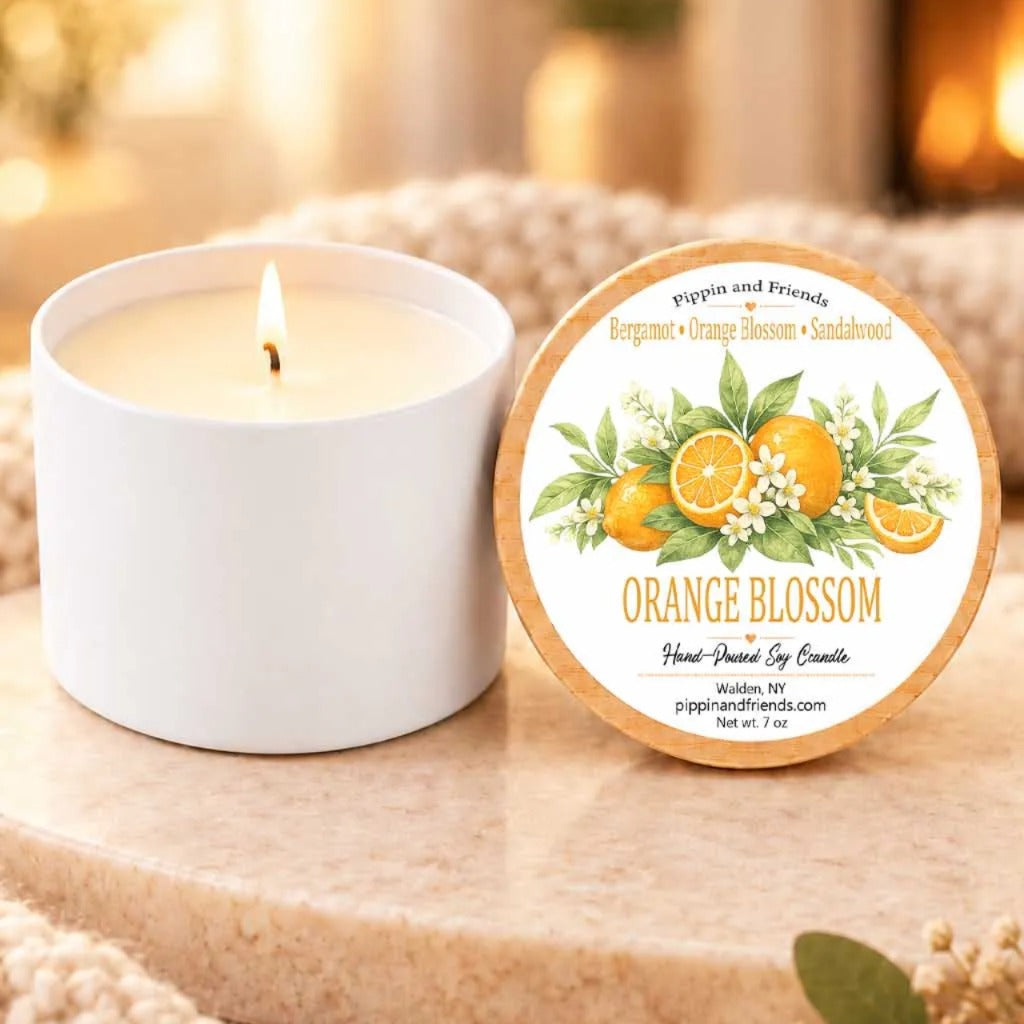a white candle with a label that reads "Orange Blossom" and "Pippin and Friends Bergamot + Orange Blossom + Sandalwood" on a table. The candle is accompanied by a small white candle holder and a few orange flowers.