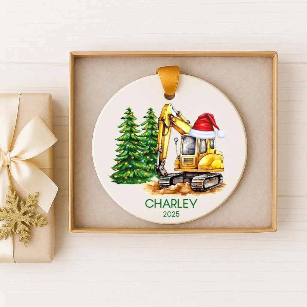 A white ornament with a yellow excavator wearing a Santa hat, surrounded by Christmas trees, is displayed in a box.