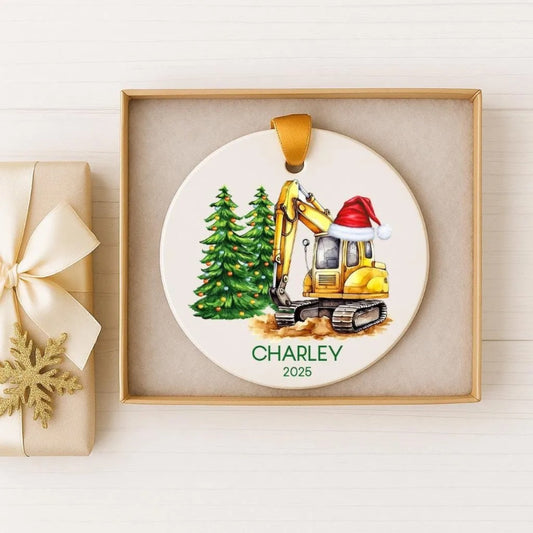 A white ornament with a yellow excavator wearing a Santa hat, surrounded by Christmas trees, is displayed in a box.