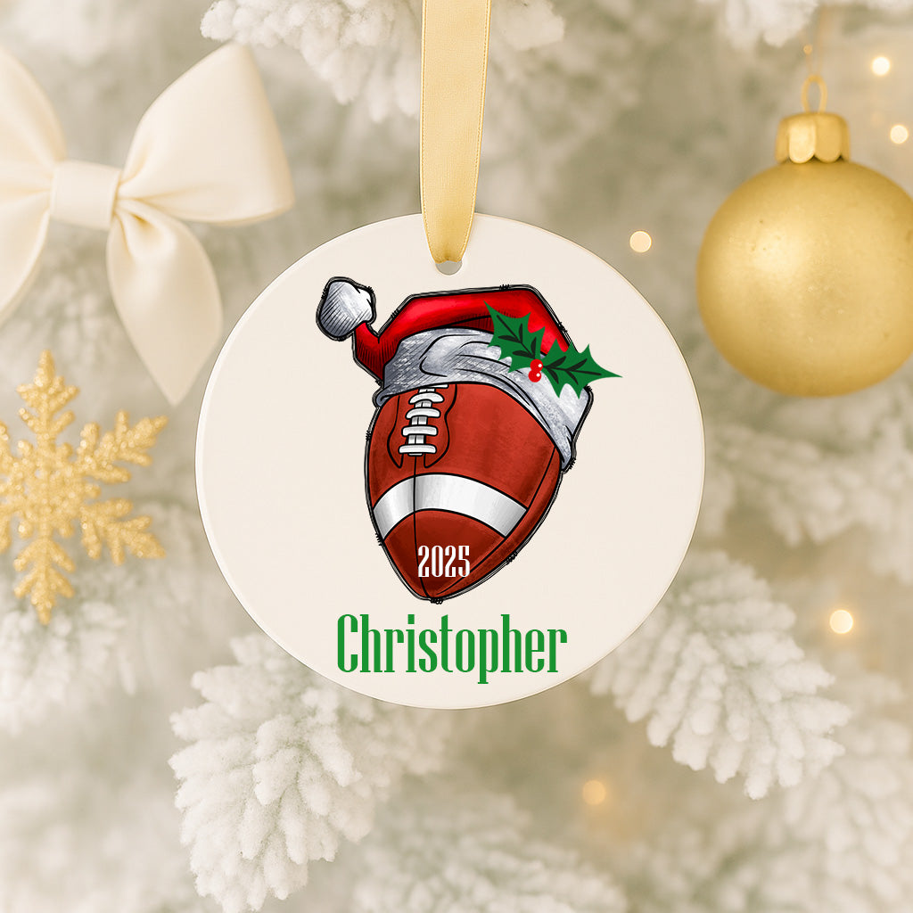 A white ornament with a red football and a Santa hat on it, hanging on a Christmas tree.