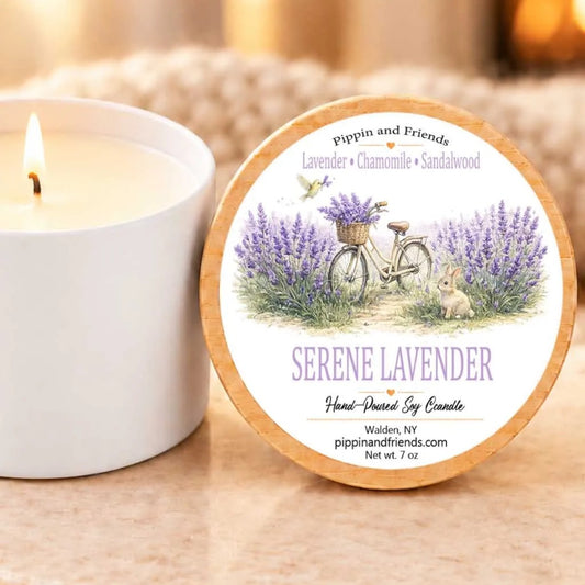 a white candle with a label that reads "Serene Lavender Hand-Dipped Soy Candle" and "Made in Walden, NY" along with the website "pippinandfriends.com" and the candle's weight "Net wt. 7 oz." The candle is placed next to a white ceramic mug, creating a cozy and inviting scene.