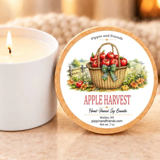 a white candle with a label that reads "Apple Harvest" and "Hand-poured by Candle" on a wooden coaster. The candle is placed next to a white candle holder, and there is a blurred background that includes a fireplace.