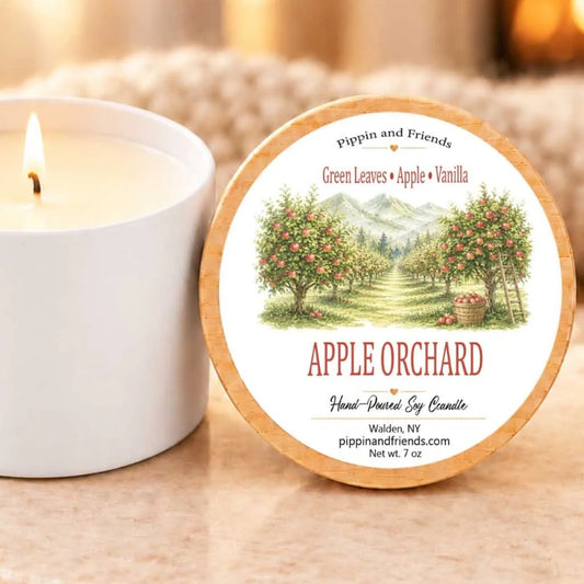 A white candle with a label that reads "Apple Orchard" sits next to a lit candle on a table.