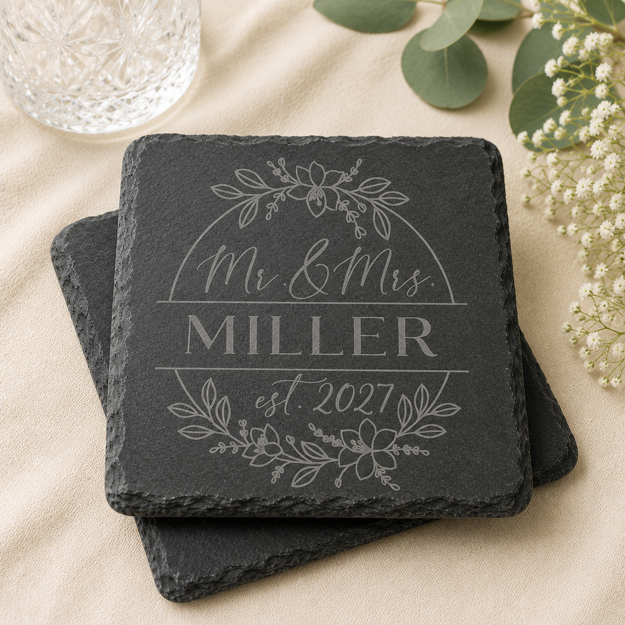 Personalized slate coaster engraved with family name and established year, displayed in an elegant wedding-inspired setting with crystal glassware and greenery.