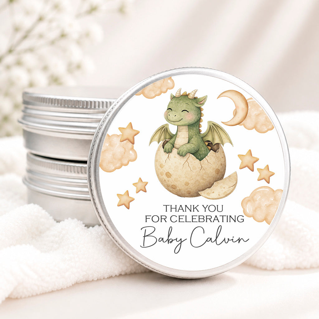 A round silver tin lid with a printed baby shower favor label featuring a green dragon hatching from an egg, stars, clouds, and the text 'THANK YOU FOR CELEBRATING Baby Calvin,' shown on top of stacked silver tins with soft white fabric and baby's breath in the background.