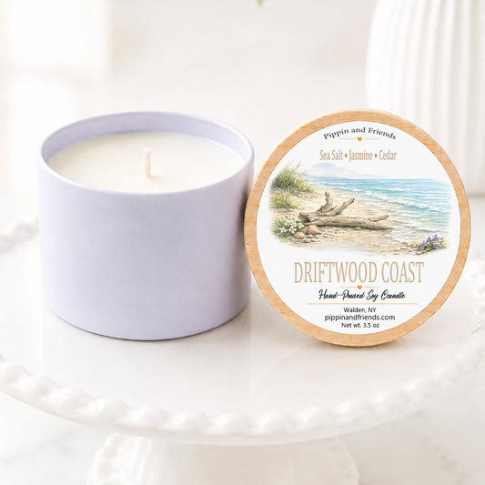 A  soy candle in a matte jar sits next to its round lid on a white pedestal. The lid features a beach illustration with the scent name 'Driftwood Coast' and the brand 'Pippin and Friends'.