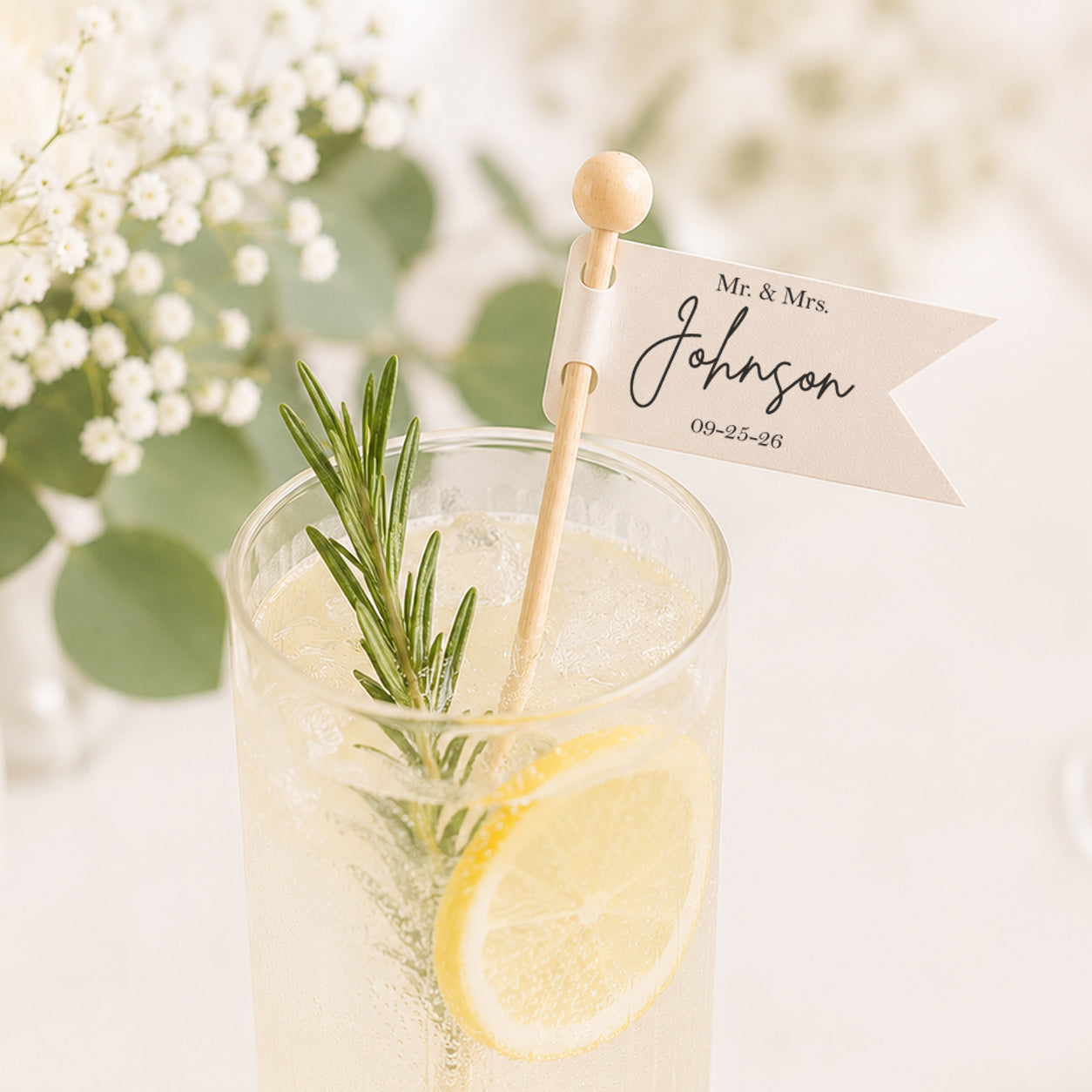 Personalized wedding drink stirrer featuring a custom ivory flag with the couple's name and wedding date attached to a 6-inch wooden stick. Displayed in a sparkling lemonade cocktail garnished with fresh rosemary and a lemon slice, styled on an elegant wedding reception table with soft florals and airy neutral décor.