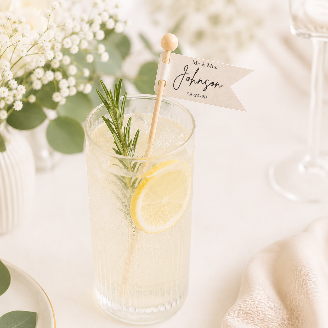 Personalized wedding drink stirrer featuring a custom ivory flag with the couple's name and wedding date attached to a 6-inch wooden stick. Displayed in a sparkling lemonade cocktail garnished with fresh rosemary and a lemon slice, styled on an elegant wedding reception table with soft florals and airy neutral décor.