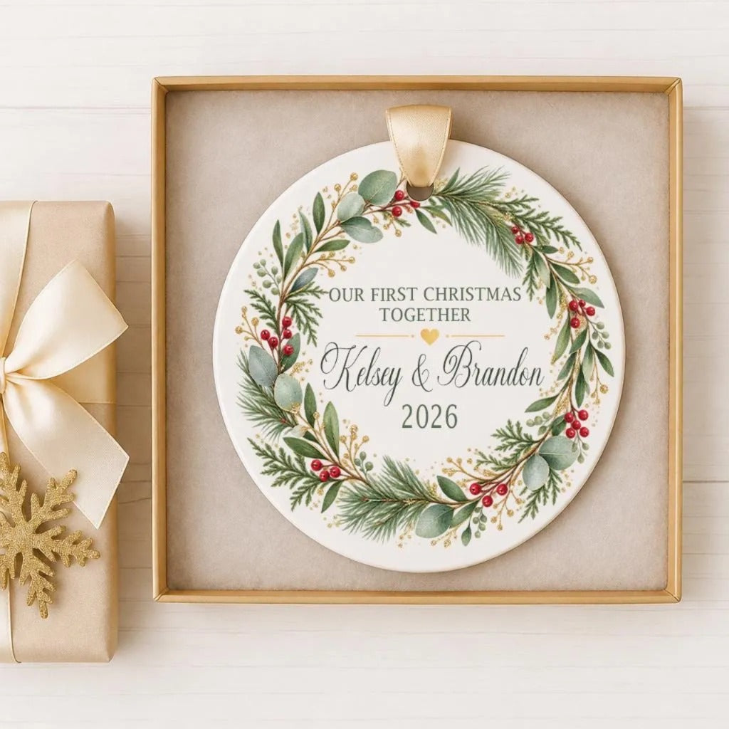 A white ceramic ornament with a wreath design and the text "Our first Christmas together Kelsey & Brandon 2026" is displayed in a wooden frame.