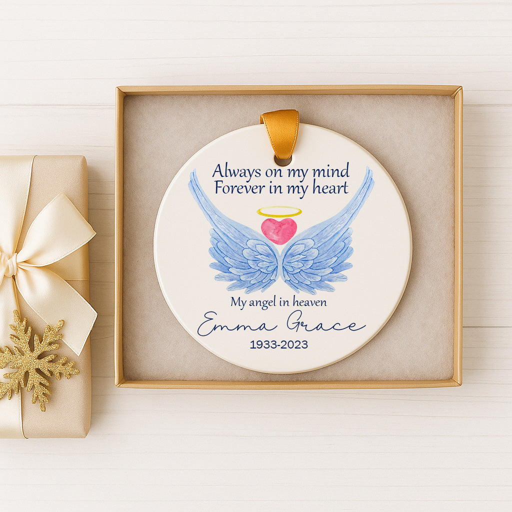 A white ceramic ornament with a blue wing design and a heart symbol, placed inside a gold box. The ornament has a message that reads "Always on my mind Forever in my heart My angel in heaven Emma Grace 1993-2023".