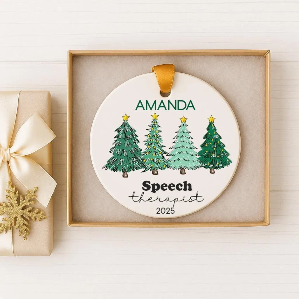 A white ornament with a gold ribbon, featuring a circular design with illustrations of four green Christmas trees and the text "AMANDA Speech Therapist 2026".