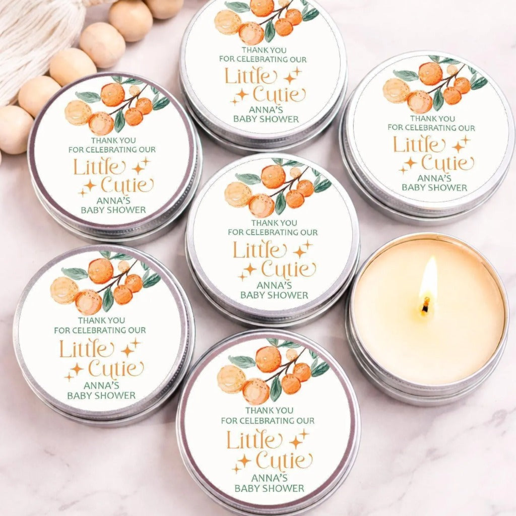 six silver tin containers with orange and green designs, arranged on a marble surface. The containers are labeled as "Little Cutie Anna's Baby Shower" and feature a candle inside.