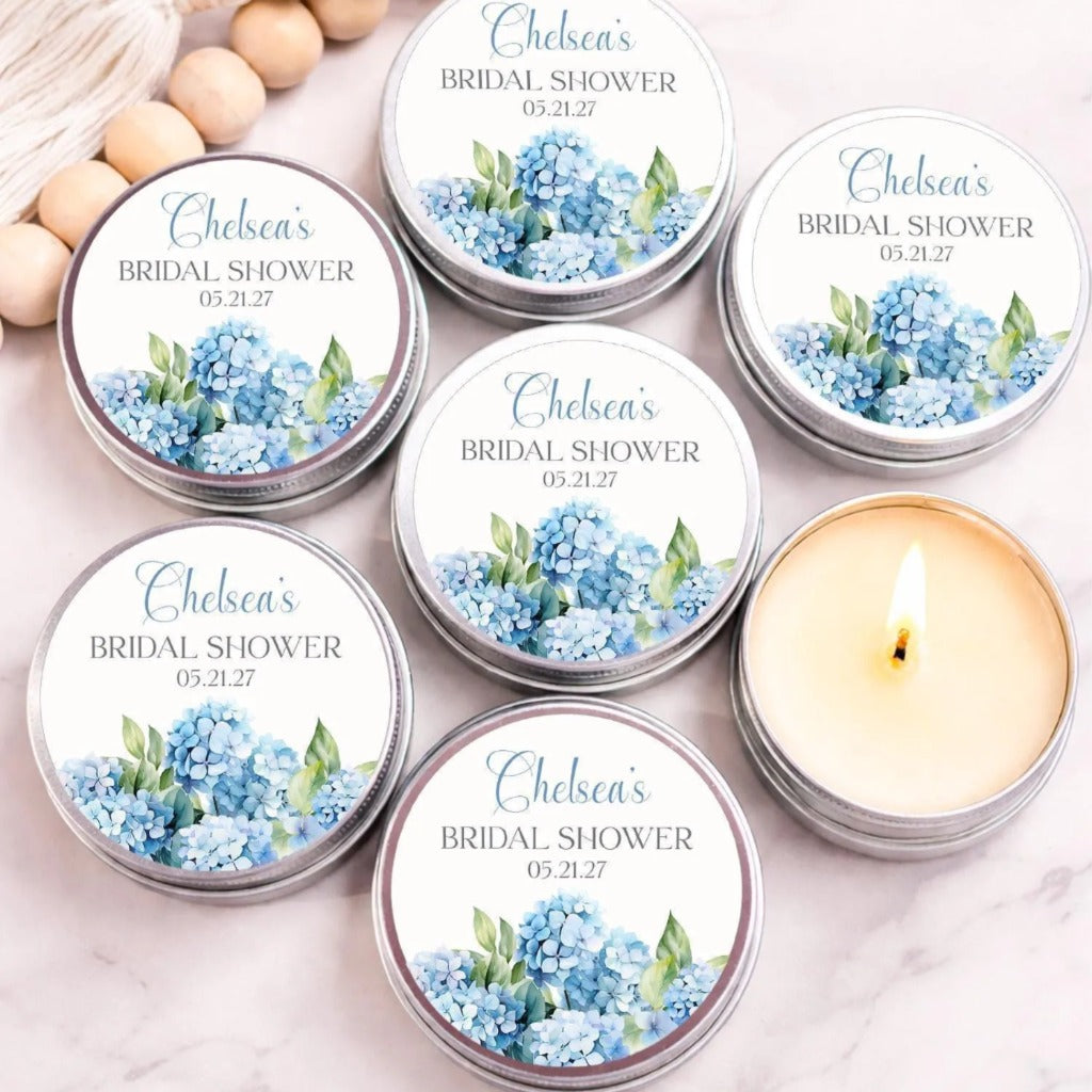 a collection of six round tin candles with labels that read "Chelsea's BRIDAL SHOWER" and "05.21.27". The candles are arranged on a marble surface, with some of them lit.