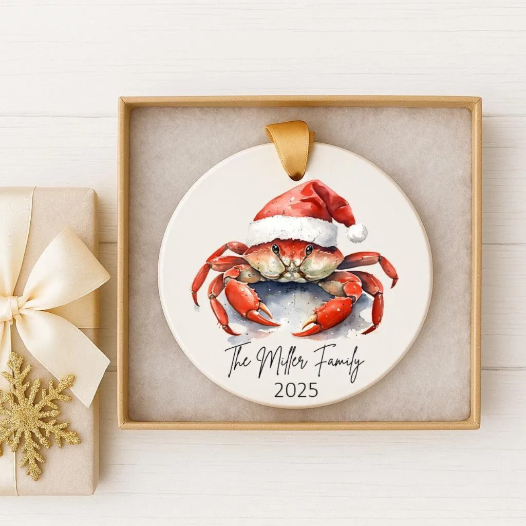 A white ornament with a red crab wearing a Santa hat, placed in a gold frame.