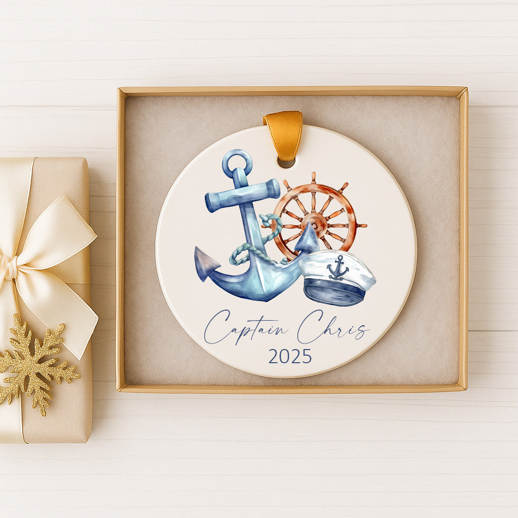 A decorative ornament featuring an anchor, a ship's wheel, and a captain's hat, with the text "Captain Chris" and the year "2025" printed on it. The ornament is displayed in a gold box, and there is a gift box with a gold ribbon next to it.