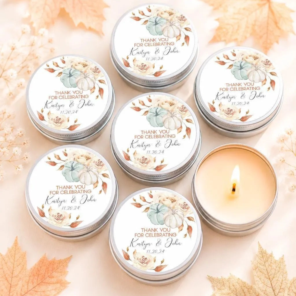a collection of six silver tins arranged on a table, each with a label that reads "Thank you for celebrating Kristyn & John 11.24.24". The tins are surrounded by autumn leaves, creating a festive and seasonal atmosphere.