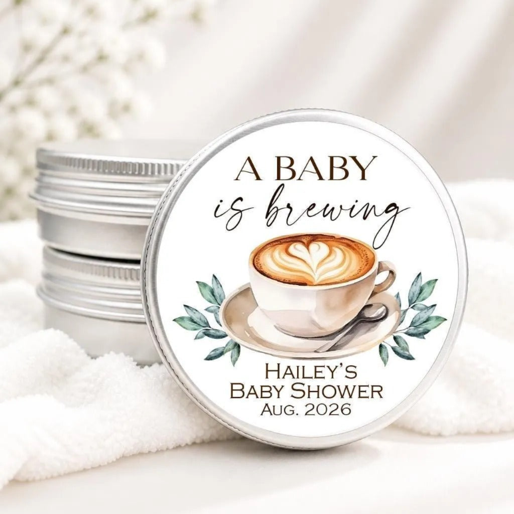 A round tin lid with a label that reads "A BABY is brewing" and features an illustration of a cup of coffee with a heart design.