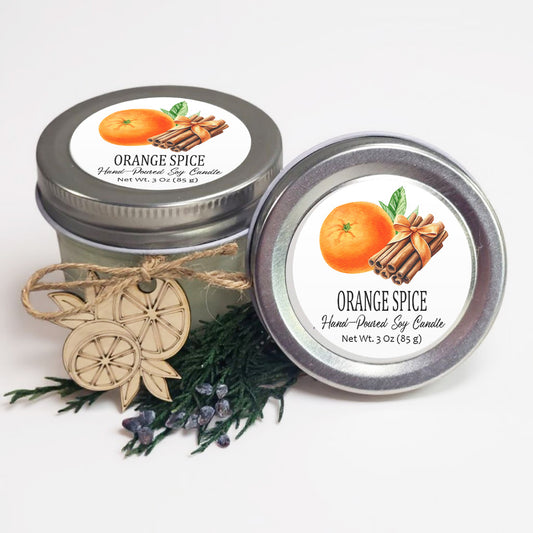 two jars of orange spice soy candles, with one jar placed on top of the other. The jars are decorated with orange and cinnamon sticks, and there are some pine branches and blueberries in the foreground.