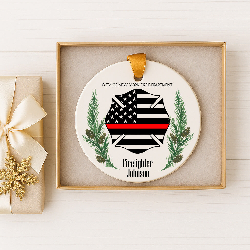 A white ceramic ornament with a black and white firefighter's helmet and a red stripe, surrounded by pine branches and a ribbon, displayed in a gold box.