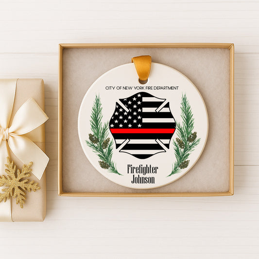 A white ceramic ornament with a black and white firefighter's helmet and a red stripe, surrounded by pine branches and a ribbon, displayed in a gold box.