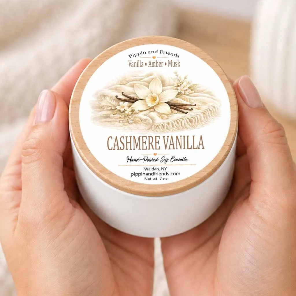 A person's hands holding a white container with a label that reads "CASHMERE VANILLA Hand-Poured Soy Candle" and features an illustration of vanilla and amber-scented flowers.