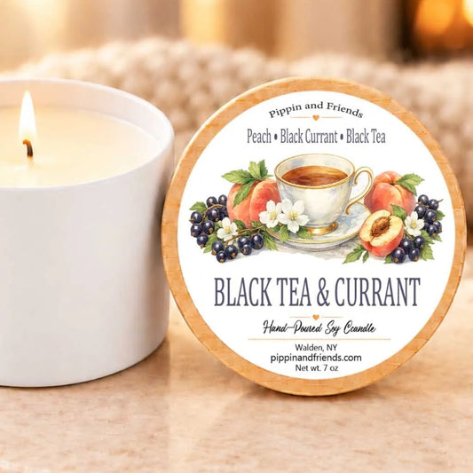 A white candle with a label that reads "Black Tea & Currant" next to it, along with a blurred background that includes a lit candle and a plant.