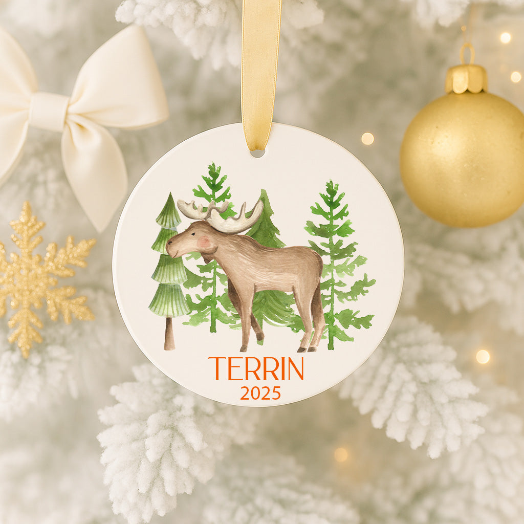 A white ornament with a brown moose and green trees, hanging on a Christmas tree.