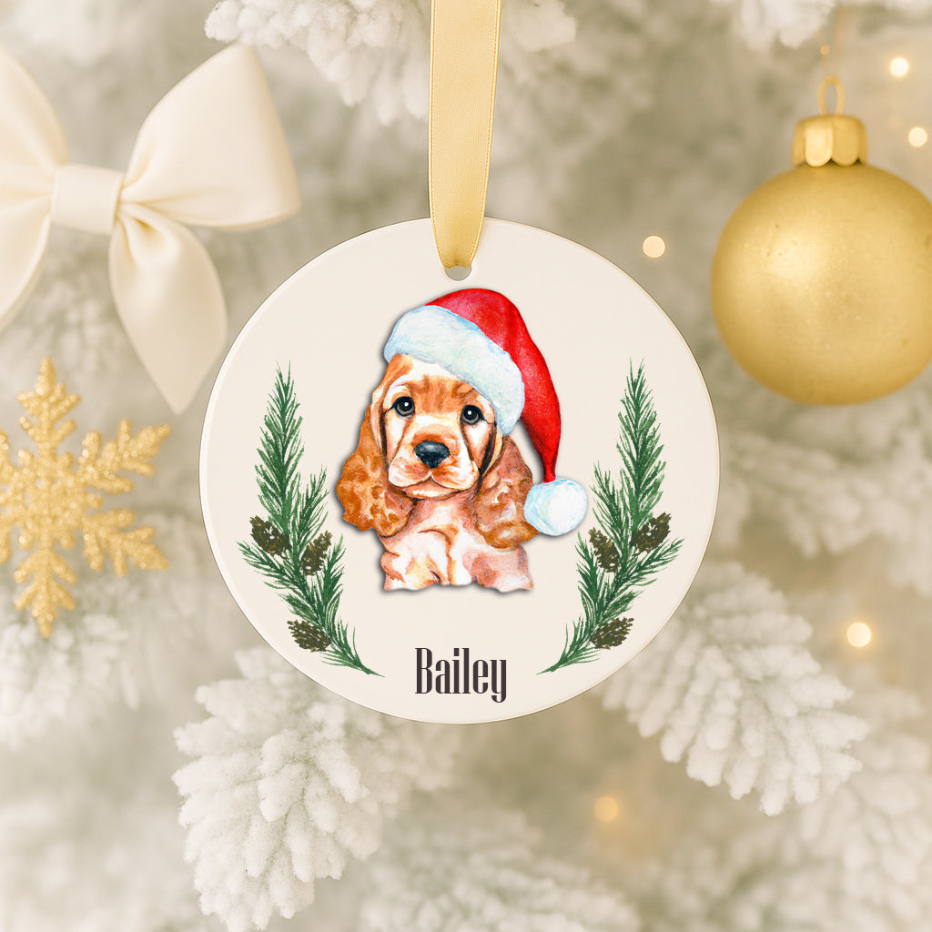 A white ornament with a picture of a dog wearing a Santa hat, surrounded by pine branches and a ribbon.