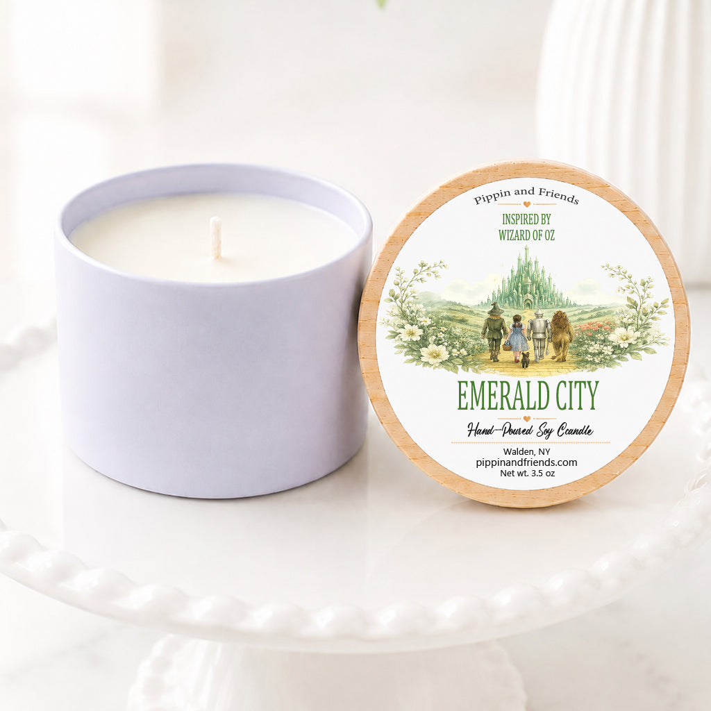 A soy candle jar sits beside its round lid on a white pedestal. The lid features a natural wood rim and a printed label reading 'Emerald City' with a small illustrated scene of people by a lake.