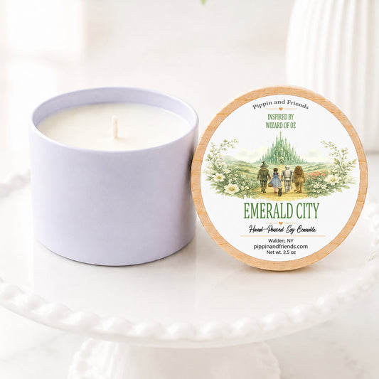 A soy candle jar sits beside its round lid on a white pedestal. The lid features a natural wood rim and a printed label reading 'Emerald City' with a small illustrated scene of people by a lake.