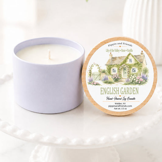 A soy candle in a matte cup container sits next to its round lid on a white pedestal. The lid features an illustration of a quaint English garden cottage with the text 'ENGLISH GARDEN' and 'Hand Poured Soy Candle' by 'pippin and friends'.