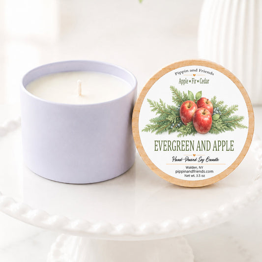 A soy candle in a matte container sits next to its round lid on a white pedestal. The lid features an illustration of red apples and green evergreen branches with the scent name 'EVERGREEN AND APPLE'.