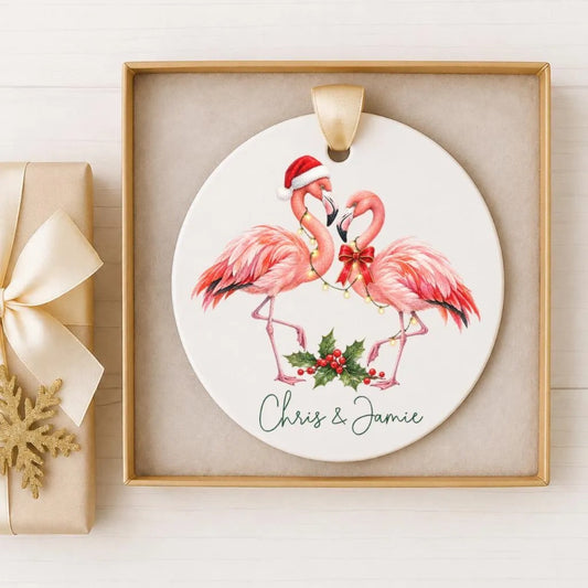A ceramic ornament featuring two pink flamingos wearing Santa hats and holding a holly leaf and berries, with the names 'Chris' and 'Jenny' written below the image.