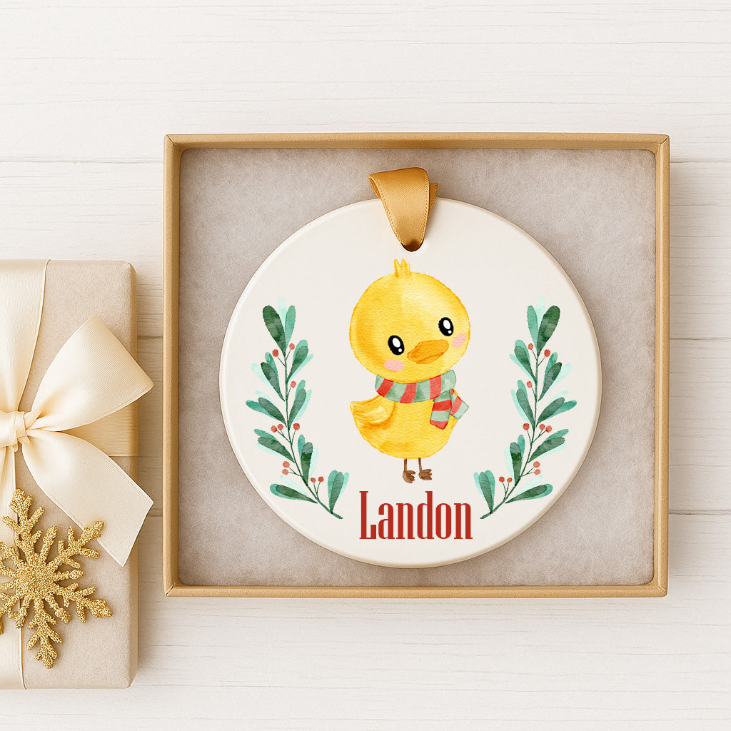 A ceramic ornament featuring a yellow duck wearing a scarf and surrounded by green leaves and berries, with the name "Landon" written on it. The ornament is displayed in a wooden frame, and there is a gift box with a white ribbon next to it.