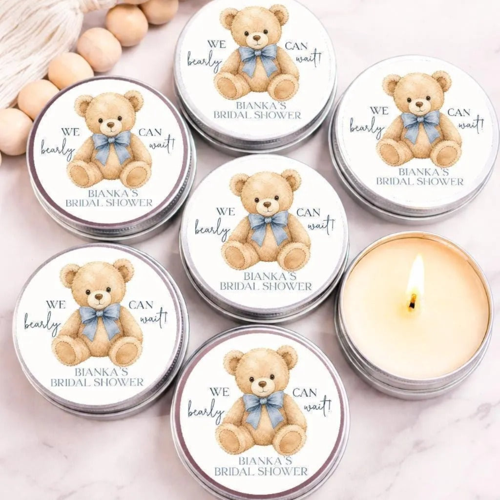a collection of round tin containers with teddy bear illustrations, arranged on a marble surface. Each container has a label that reads "We can wait for BANKA's BRIDAL SHOWER" and features a teddy bear with a blue bow. There is also a single lit candle in the center of the arrangement.