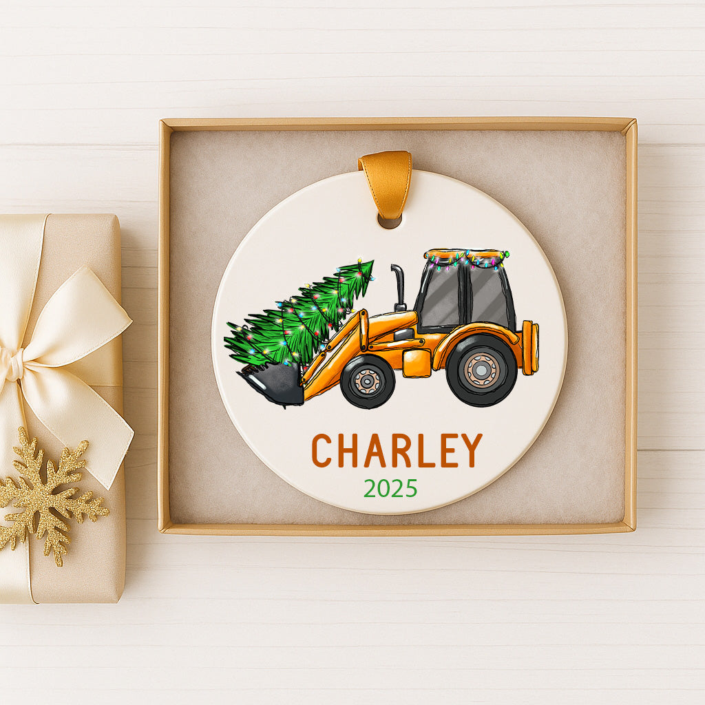 A Christmas ornament in a box, featuring a tractor with a Christmas tree in its bed.