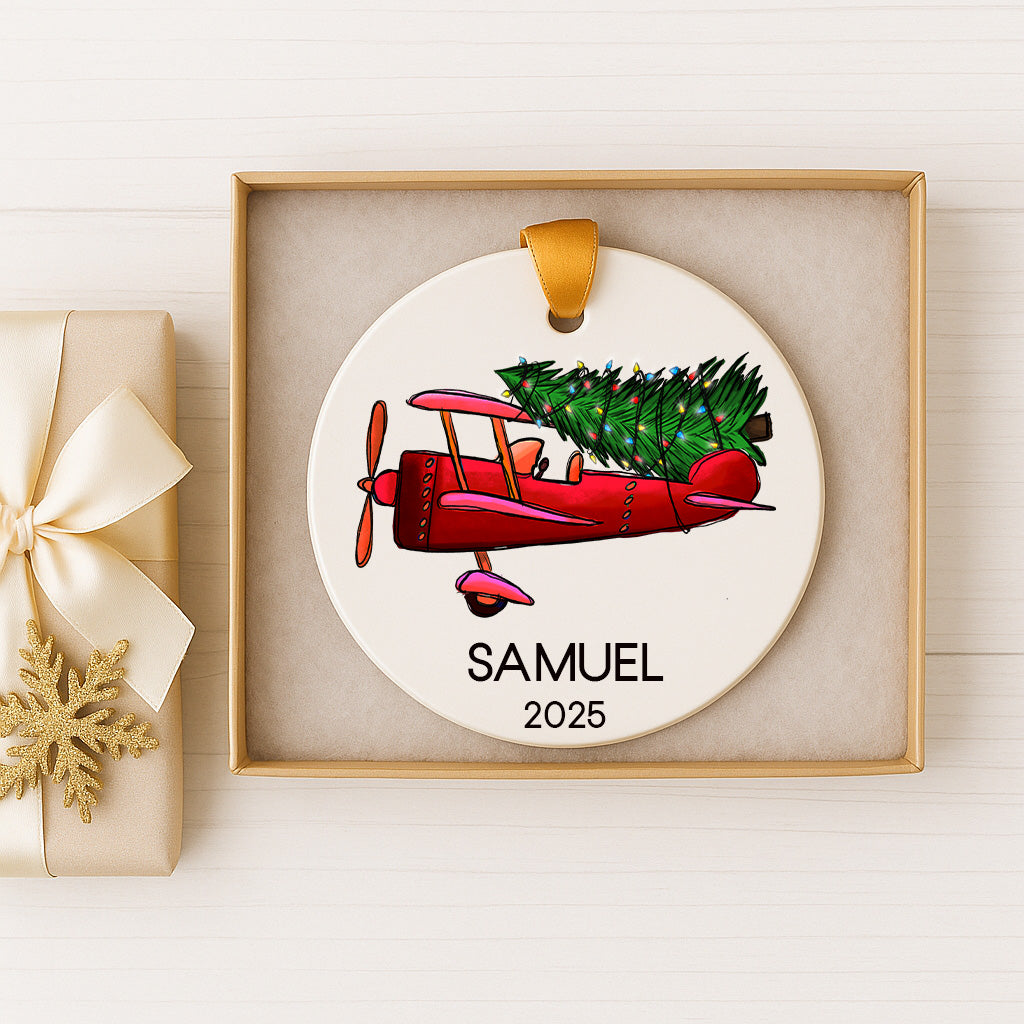 A white ornament with a red airplane and a Christmas tree, displayed in a gold box.