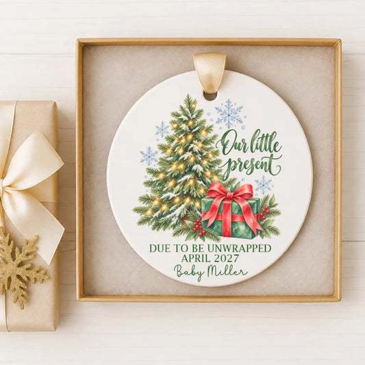 A white ceramic ornament with a Christmas tree and a red bow, hanging on a wooden surface. The ornament has text that reads "Our little present Due to be unwrapped April 2027 Baby Miller".