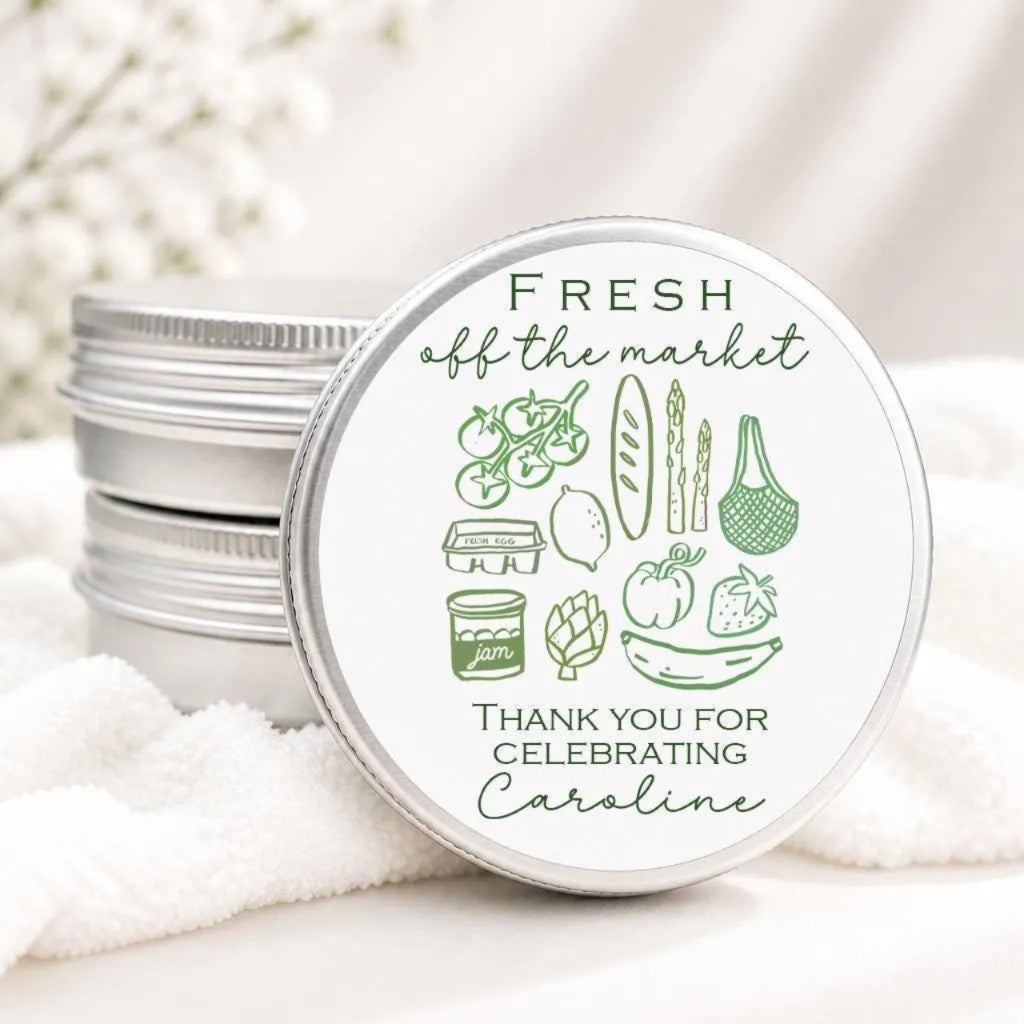 a round tin lid with a green and white design, which includes various food items such as tomatoes, bread, and bananas, along with the text "FRESH off the market Thank you for celebrating Caroline".