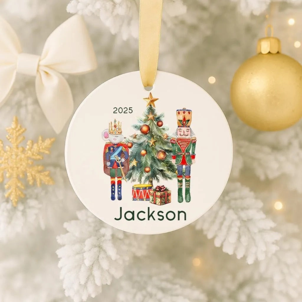 A white ornament with a Christmas tree and nutcracker characters, along with a gold ornament, is hanging on a tree.