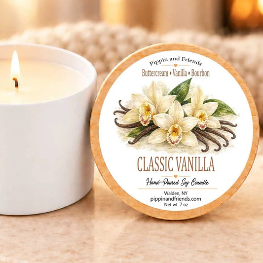 a white candle with a label that reads "Classic Vanilla" and "Hand-Poured by Candle" on a marble surface. Next to the candle is a white metal tin with a lit candle inside.