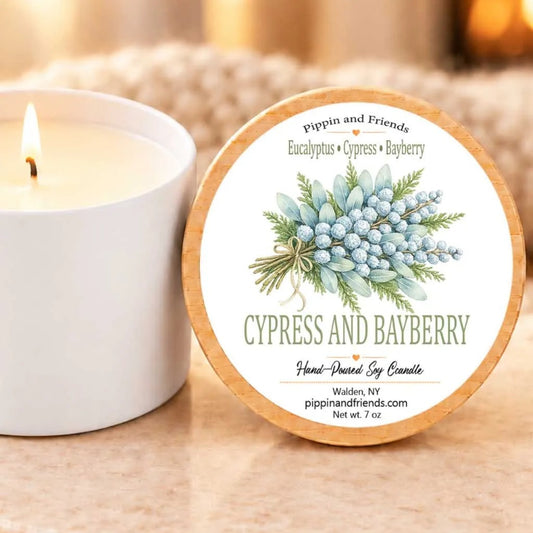 a white candle with a label that reads "Cypress and Bayberry" and "Hand-poured by Candle" on a marble coaster. The candle lid is next to a white tin, and there is a blurred background that includes a fireplace.