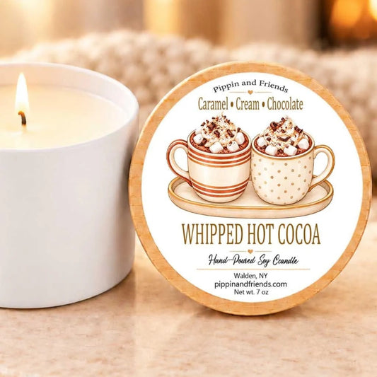 A white candle with a label that reads "Whipped Hot Cocoa" next to a candle in a white holder.