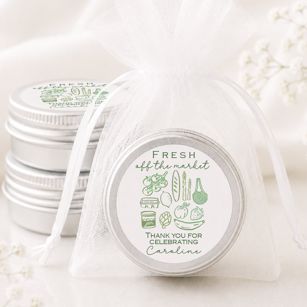 A small round silver tin wrapped in a sheer white organza drawstring bag, with a printed circular label reading 'FRESH off the market' and 'THANK YOU FOR CELEBRATING Carolina.' Additional matching silver tins are stacked in the background on a soft white fabric surface with delicate floral props.