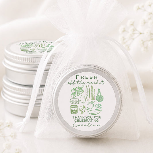 A small round silver tin wrapped in a sheer white organza drawstring bag, with a printed circular label reading 'FRESH off the market' and 'THANK YOU FOR CELEBRATING Carolina.' Additional matching silver tins are stacked in the background on a soft white fabric surface with delicate floral props.