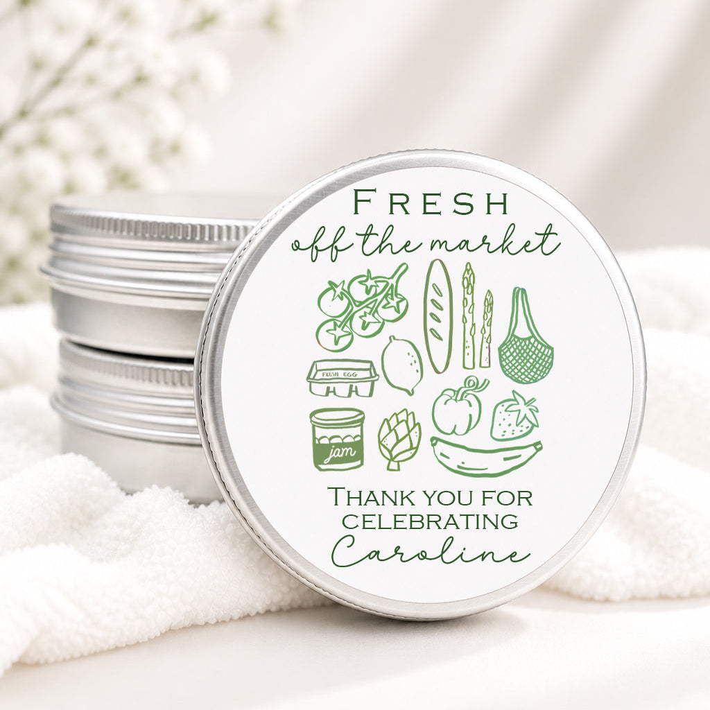 A round silver metal tin lid with green illustrated grocery items and the text 'FRESH off the market' and 'THANK YOU FOR CELEBRATING Caroline,' shown resting on white fabric with stacked matching tins behind it.