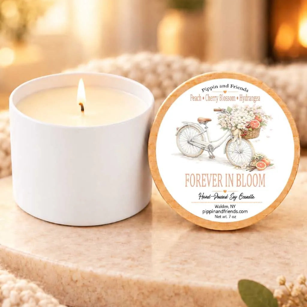 a white candle with a label that reads "Forever in Bloom" and a bicycle illustration, placed on a wooden surface next to a white candle holder.