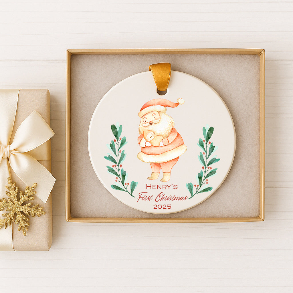A ceramic ornament in a gold box, featuring a Santa Claus figure holding a baby. The ornament has a label that reads "Henry's First Christmas 2025".