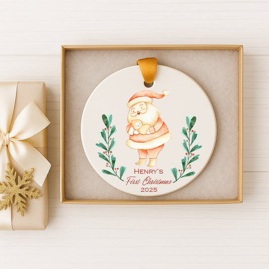 A ceramic ornament in a gold box, featuring a Santa Claus figure holding a baby. The ornament has a label that reads "Henry's First Christmas 2025".